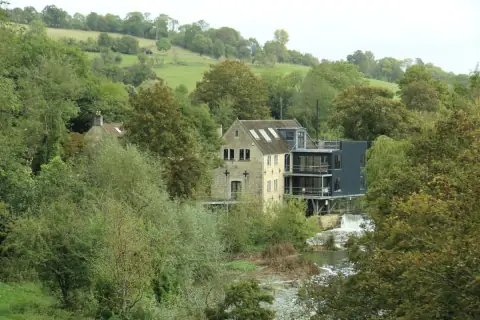 An old stone mill with a modern glass extension beside a river, among trees and fields