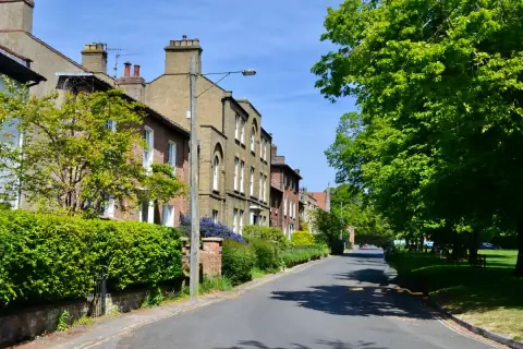 A sunny street of Victorian brick houses with hedges, opposite a green lined with trees