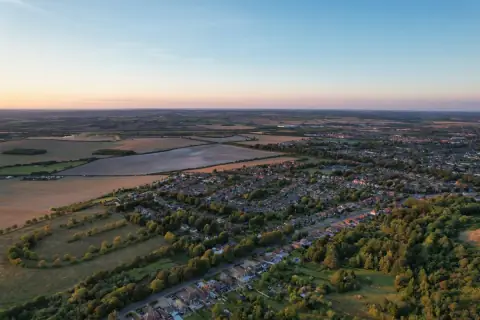 Aerial view of the edge of a town giving way to open farmland at sunset, the kind of land promoted for development